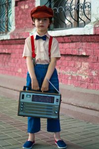 Young boy in retro fashion holding a vintage radio against a brick wall backdrop. Outdoors scene.